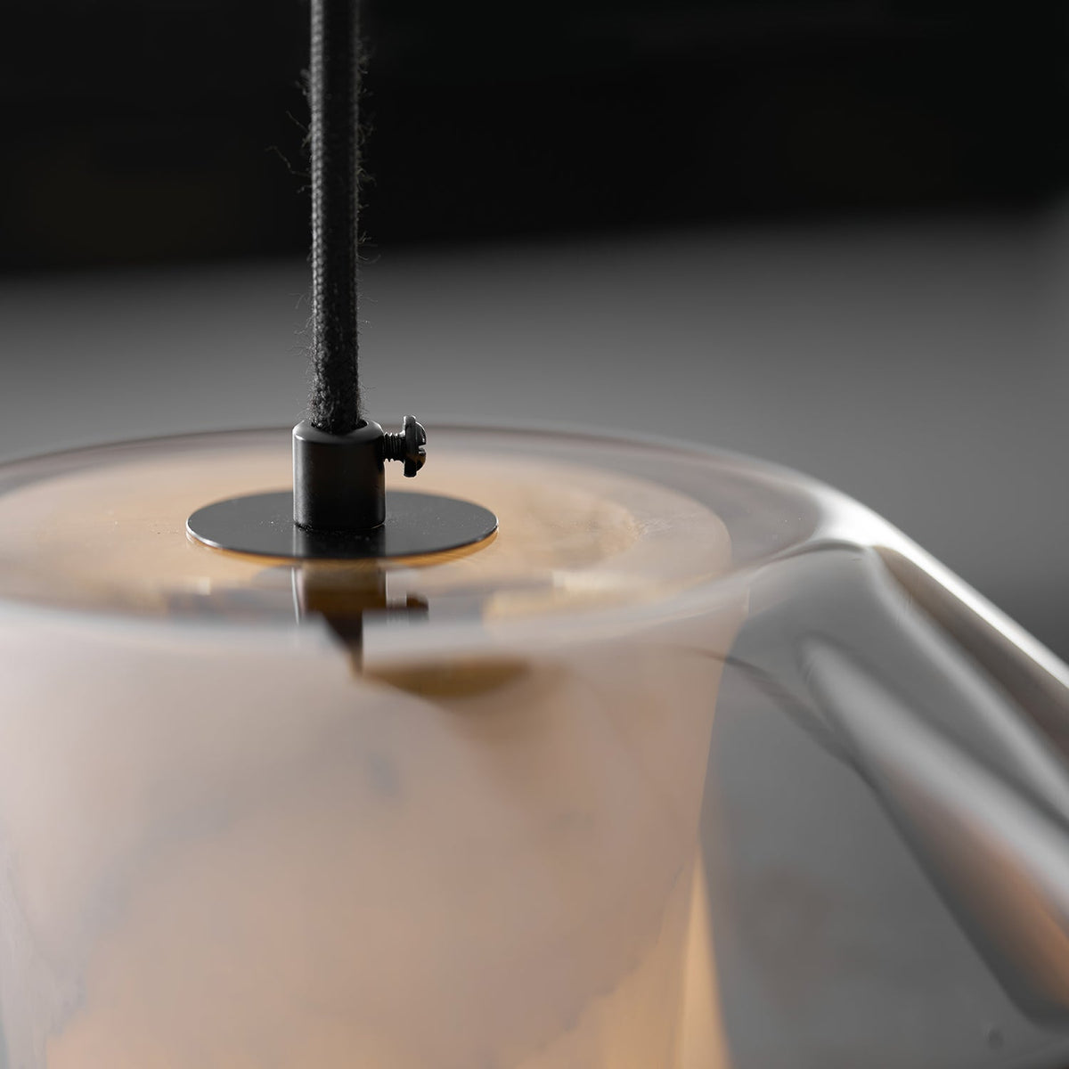 Very close view of the black cord grip and brass-toned disc at the top of this Clérachic pendant, with the alabaster glowing below.