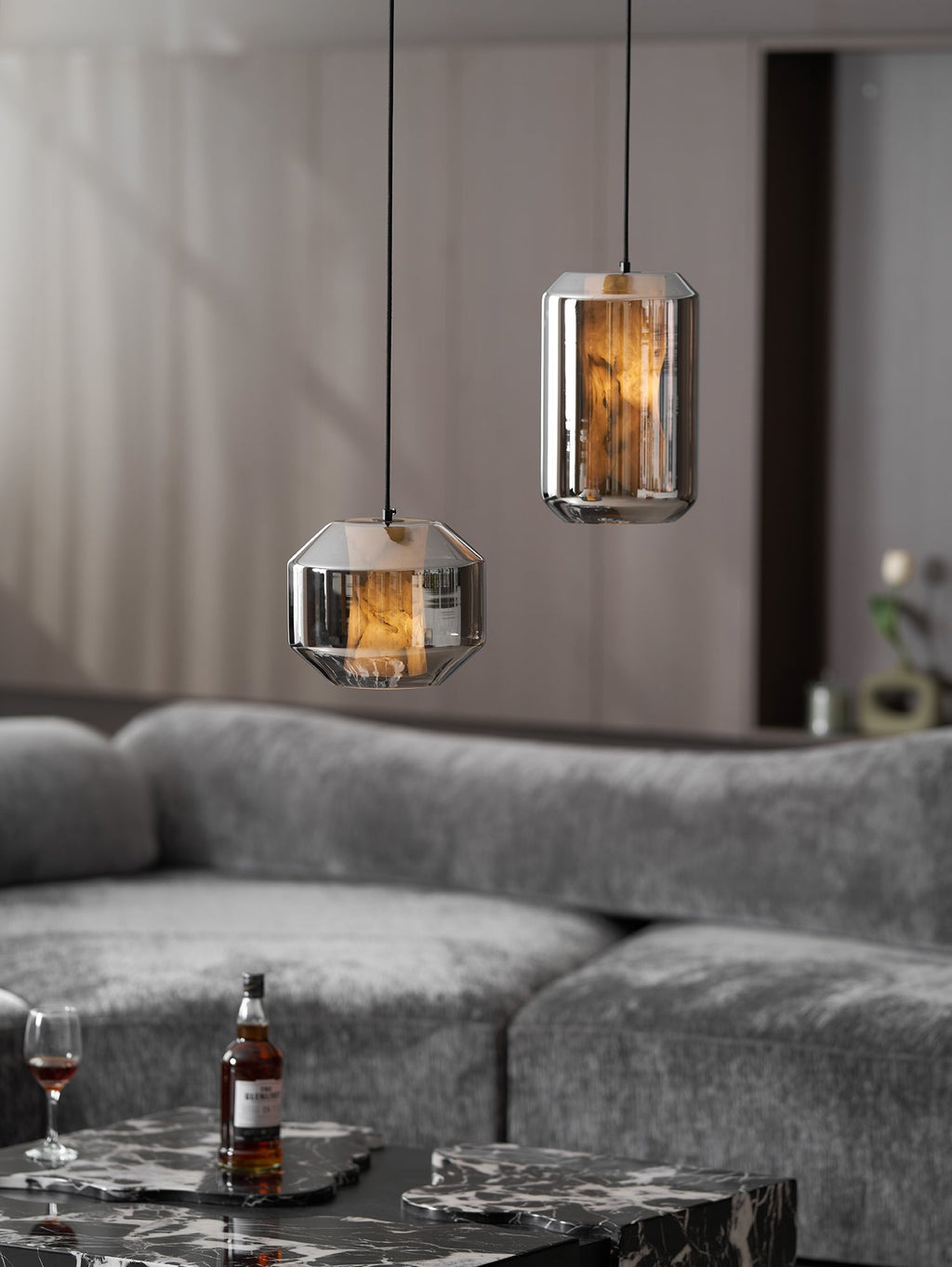 Two smoke grey Clérachic pendants hang above a grey sofa, with a bottle and a glass of whisky on a dark marble table below.