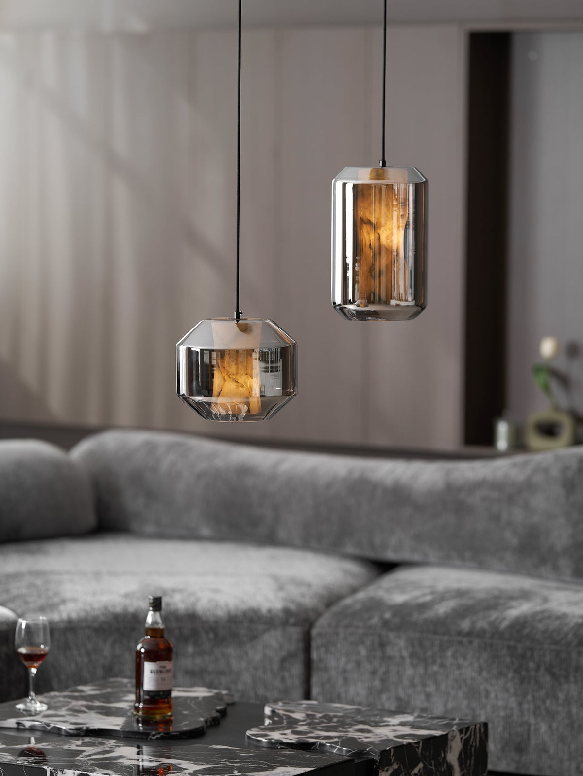 Two smoke grey Clérachic pendants hang above a grey sofa, with a bottle and a glass of whisky on a dark marble table below.