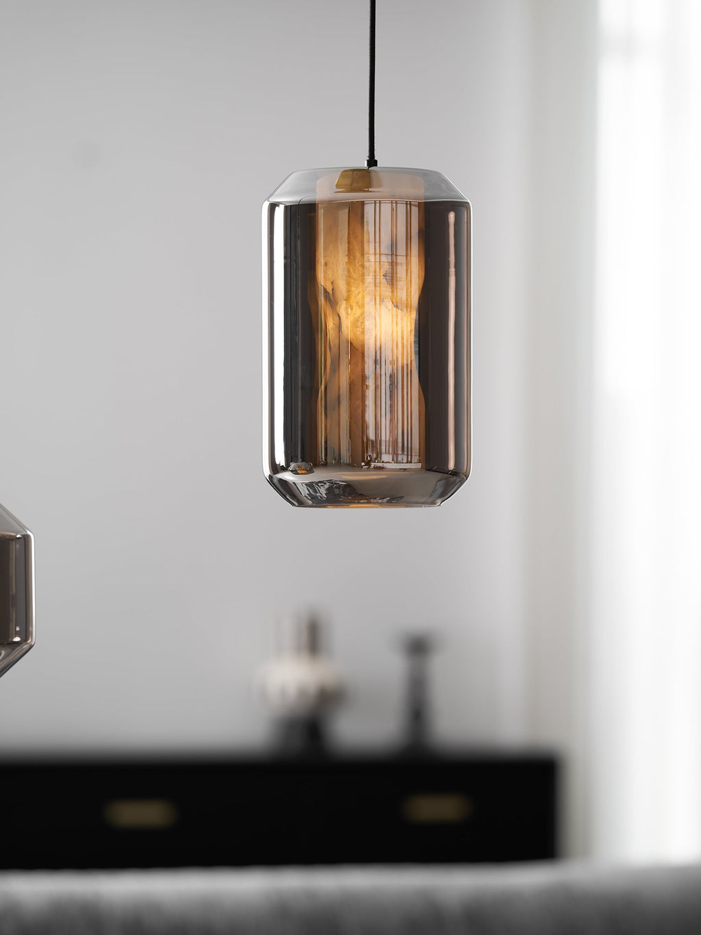 A tall smoke grey glass Clérachic pendant glows amber in front of a blurred black sideboard with brass handles and a pale curtain.