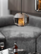 A wide smoke grey glass Clérachic pendant glows amber over a grey sofa, with a whisky glass and bottle softly out of focus below.