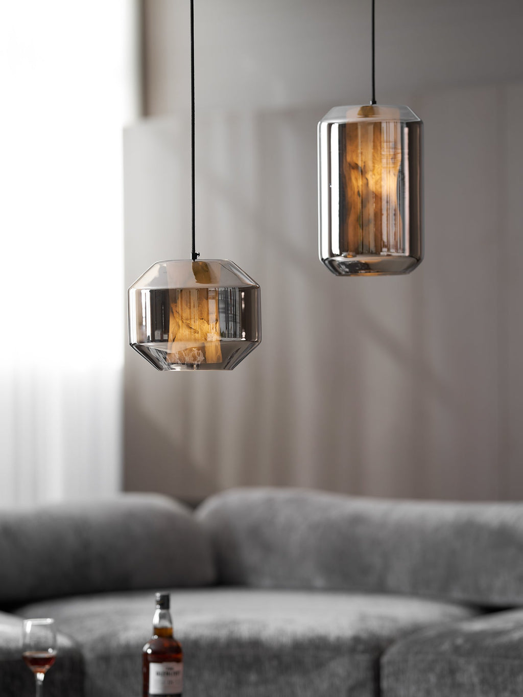 A tall and a wide smoke grey Clérachic pendant hang at different heights above a grey sofa, the tall one lit amber and the wide one dimmer.