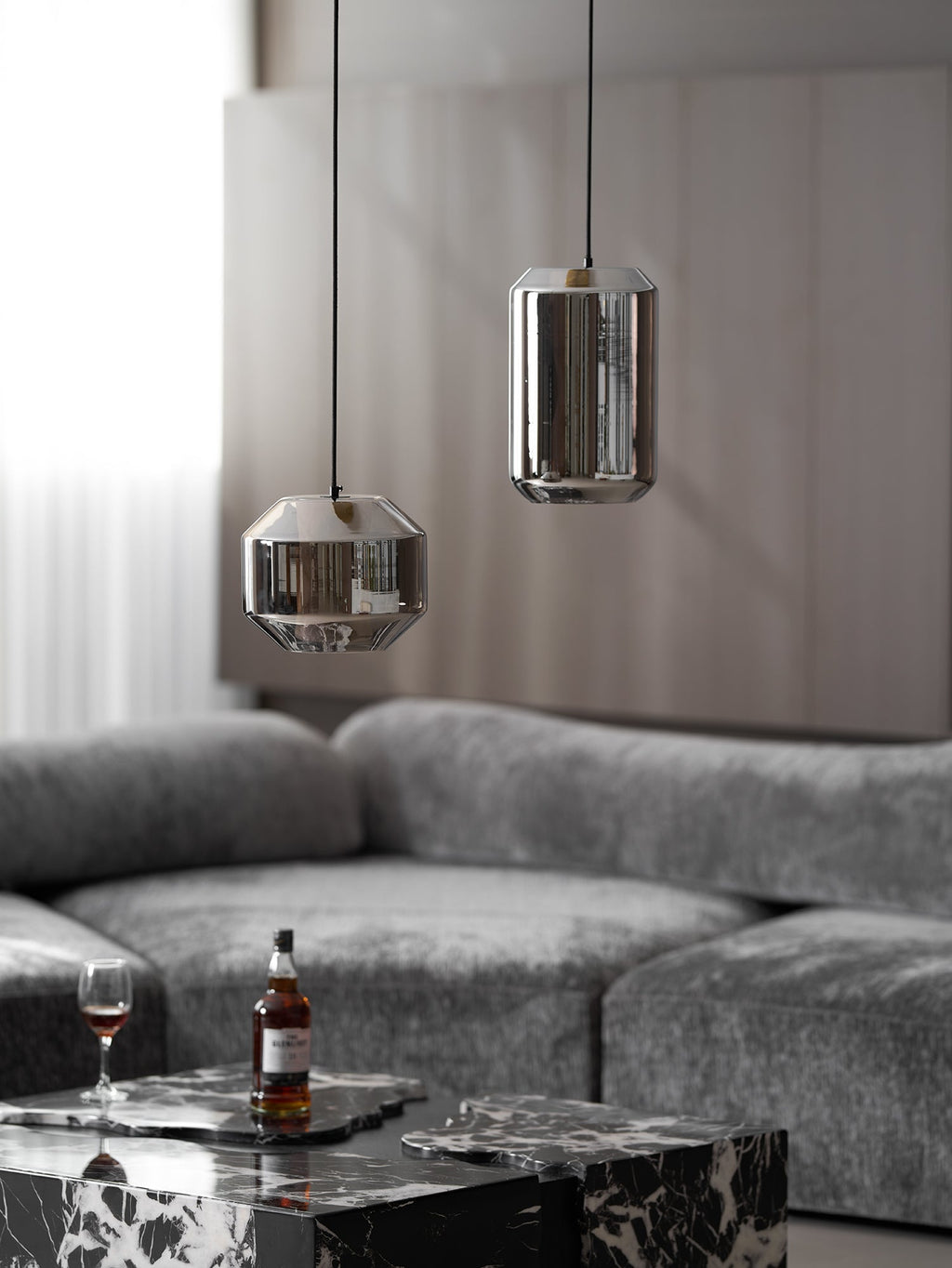 A wide smoke grey Clérachic pendant and a tall unlit one hang above a grey sofa, with a bottle and glass on a marble table below.