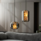 Close view of a wide and a tall smoke grey Clérachic pendant glowing amber in a dim room, with a grey sofa softly blurred below.