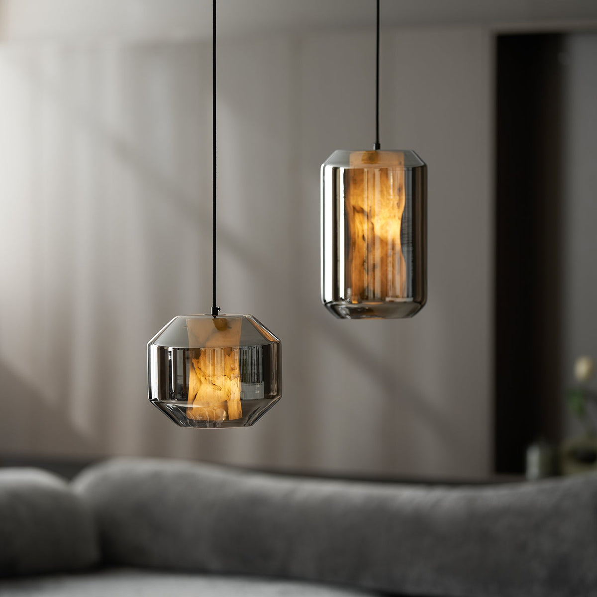 Close view of a wide and a tall smoke grey Clérachic pendant glowing amber in a dim room, with a grey sofa softly blurred below.