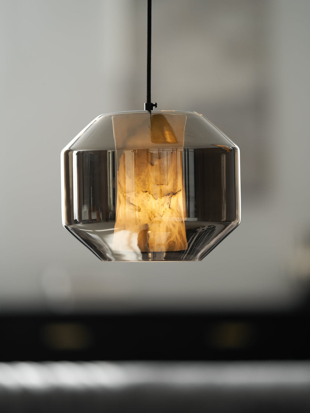 A wide smoke grey glass Clérachic pendant with its alabaster glowing amber, against a dark blurred interior.