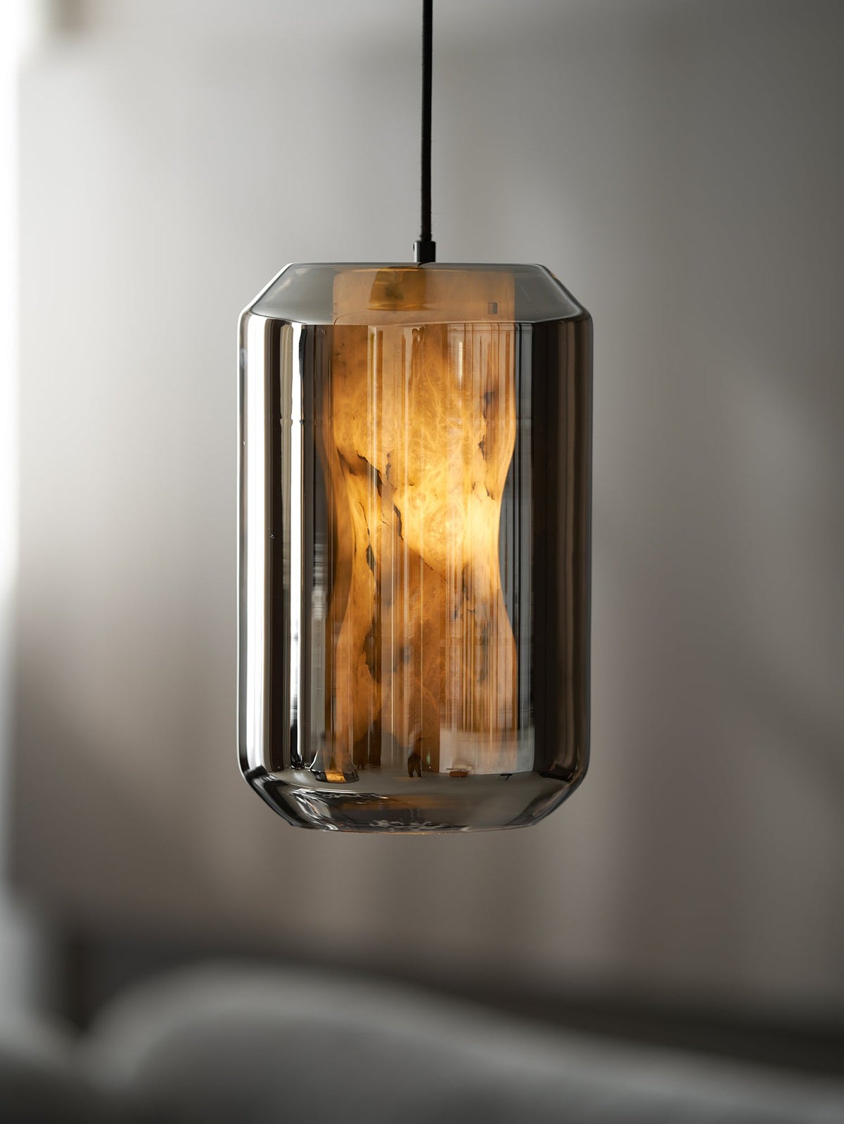 A tall smoke grey glass Clérachic pendant with its waisted alabaster column glowing amber, against a dim, softly blurred room.