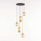 Five clear glass Clérachic pendants in wide and tall shapes hang at staggered heights from one round black canopy on a white backdrop, each lit amber.