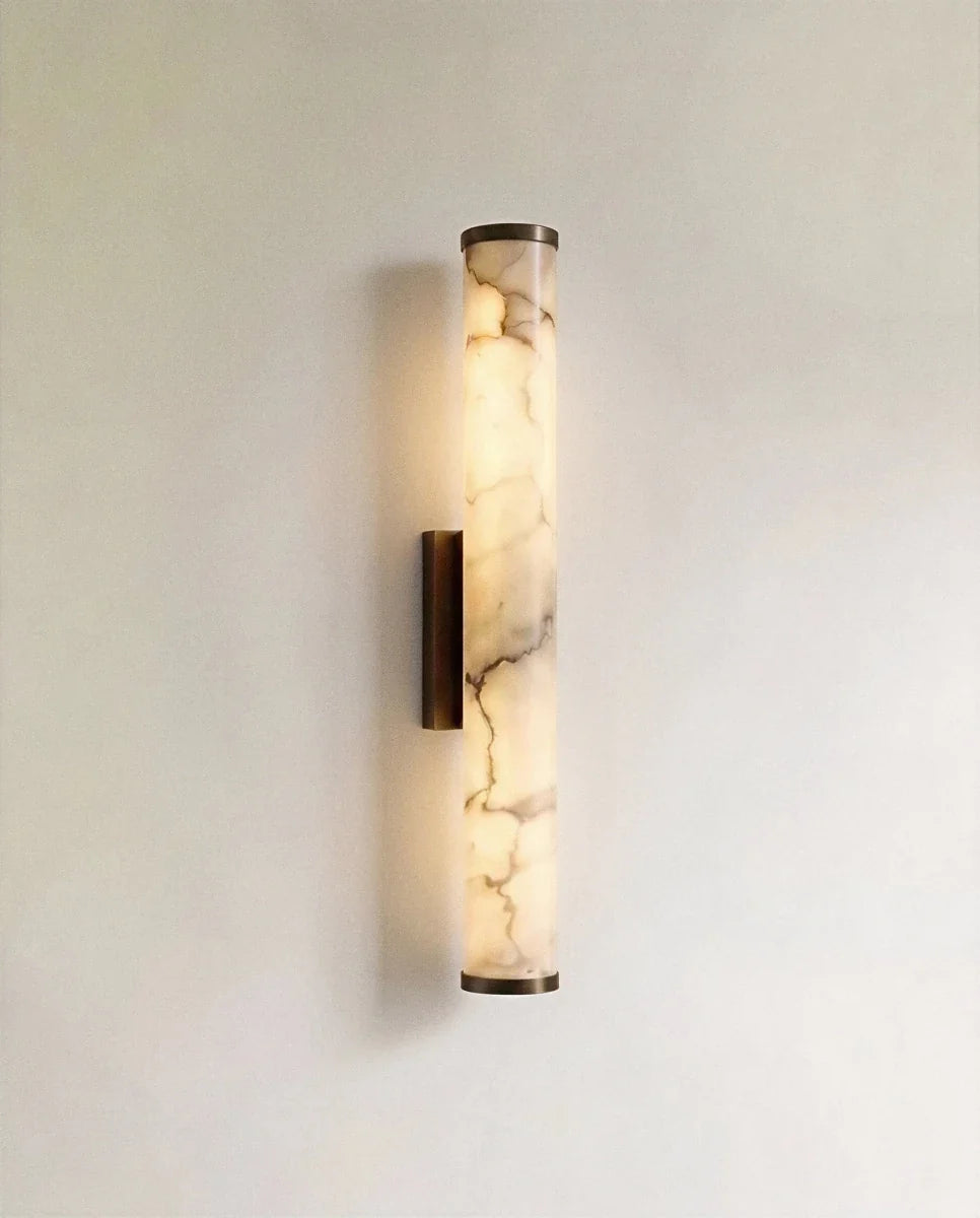 Clérachic wall light with a slender alabaster cylinder glowing warm, veining visible, on a dark metal backplate against a pale wall.