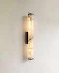 Clérachic wall light with a slender alabaster cylinder glowing warm, veining visible, on a dark metal backplate against a pale wall.