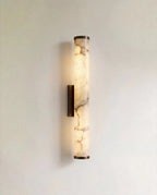 Clérachic wall light with a slender alabaster cylinder glowing warm, veining visible, on a dark metal backplate against a pale wall.