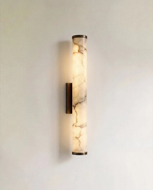 Clérachic wall light with a slender alabaster cylinder glowing warm, veining visible, on a dark metal backplate against a pale wall.