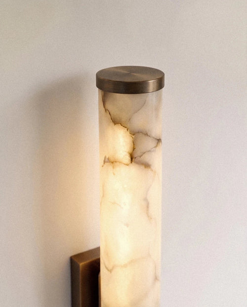 Close view of the Clérachic alabaster wall light's dark metal end cap and glowing cylinder, cloudy veining lit from within.