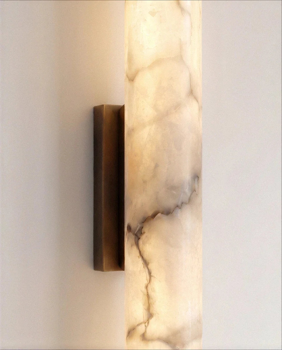 Clérachic wall light close-up showing the rectangular dark metal backplate holding a lit alabaster cylinder with brown veining.