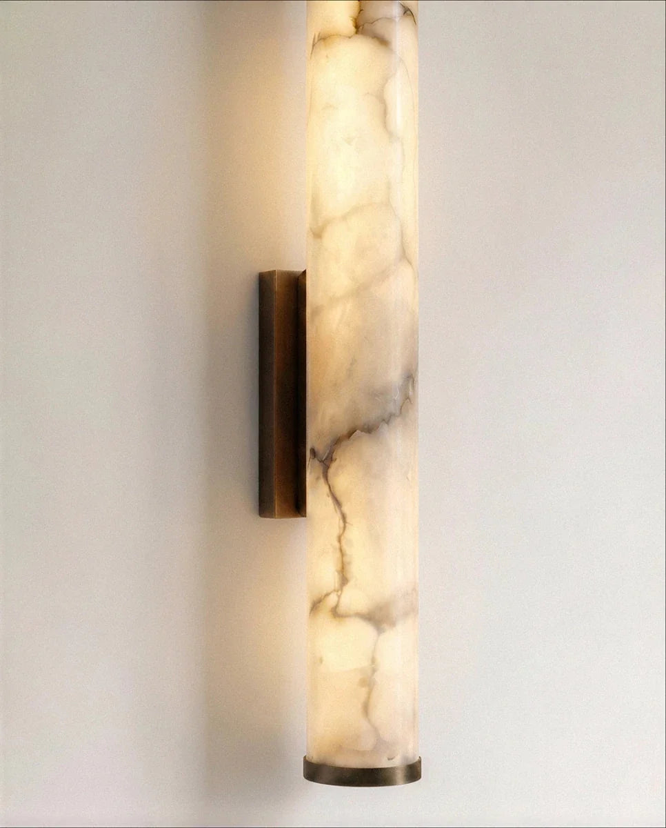 Clérachic alabaster cylinder wall light lit warm, with a dark metal wall bracket and bottom cap, veining in soft grey and amber.