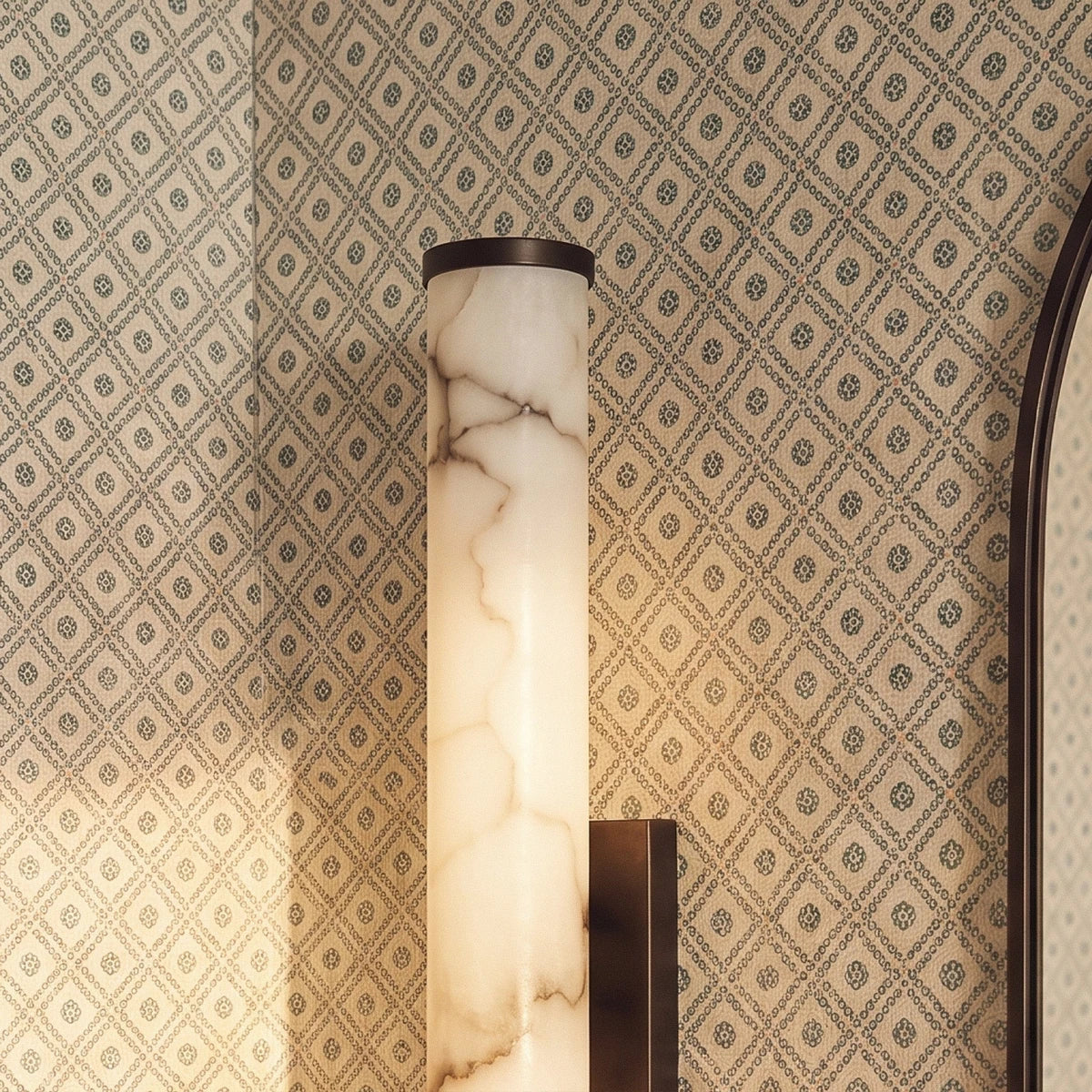 Clérachic alabaster cylinder wall light with a dark metal cap, glowing against green and cream patterned wallpaper.