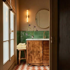 Clérachic alabaster wall light above a vanity with a round brass mirror, green wall tiles and a chequered terracotta floor.