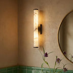Clérachic alabaster cylinder wall light lit warm on a textured plaster wall beside a round brass-framed mirror and green tiles.