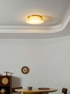 Small, warmly lit Clérachic disc light on a white ceiling with a curved moulding, a round table and a cane cabinet below.