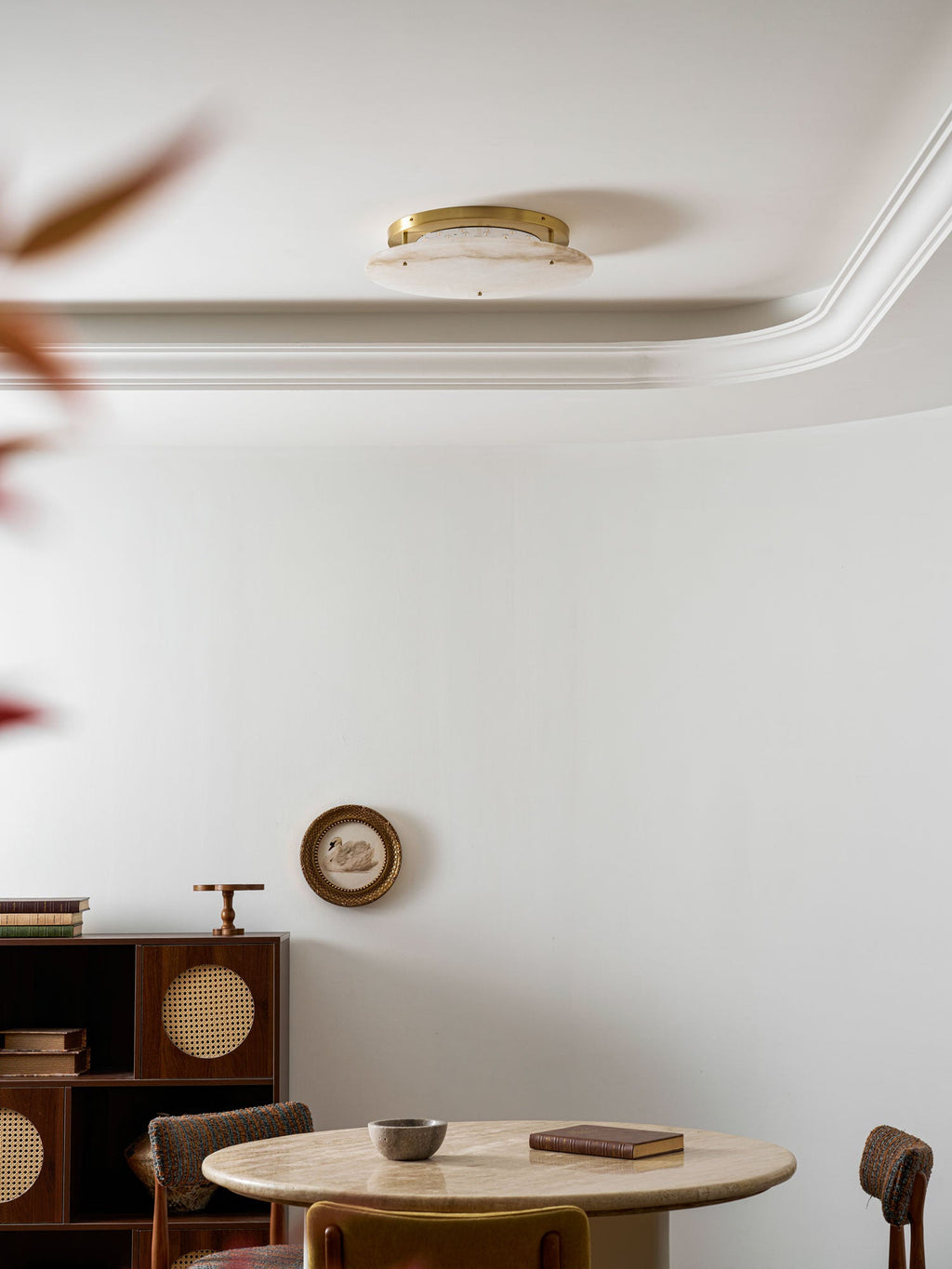 Unlit Clérachic alabaster disc high on a white ceiling by a curved moulding, with a dark red leaf at the edge of the frame.