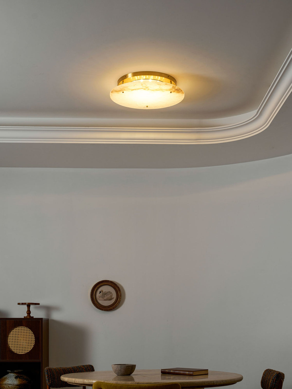 Clérachic disc light glowing warm gold under a ceiling moulding, above a round stone table with a book and a cup.