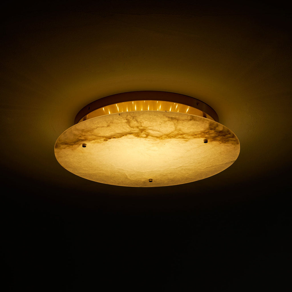 Clérachic alabaster disc light turned on in a dark room, showing the brass canopy and a row of small lights behind the stone.