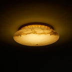 Clérachic alabaster disc light turned on in a dark room, showing the brass canopy and a row of small lights behind the stone.