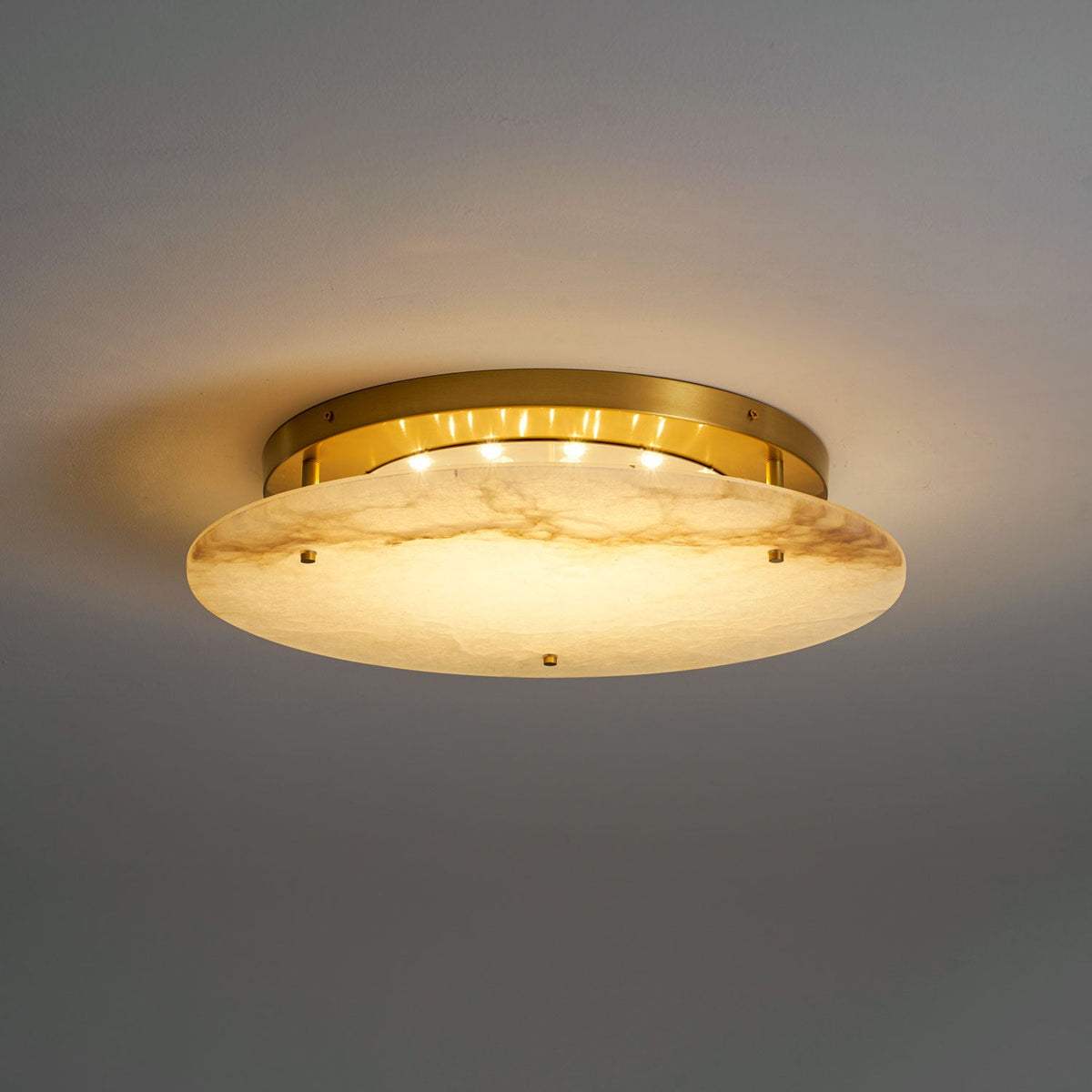 Lit Clérachic disc light on a grey ceiling, its brass canopy and small LED points glowing above the alabaster.