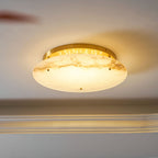 Side view of the Clérachic alabaster disc switched on, its brass ring glowing above a ceiling edge and a glass shelf below.