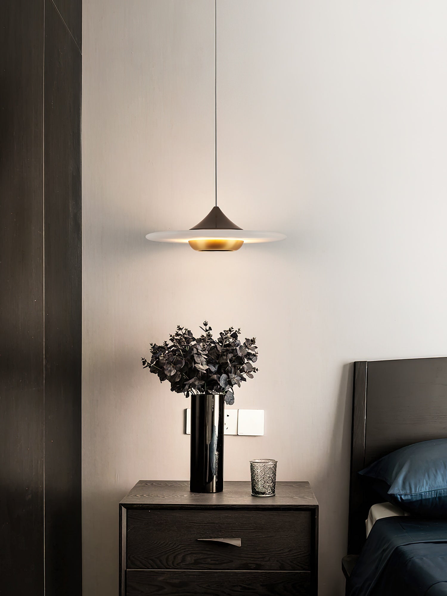 The Clérachic Alabaster Disc Pendant Light lighting a bedside chest topped with dried eucalyptus in a tall black vase.