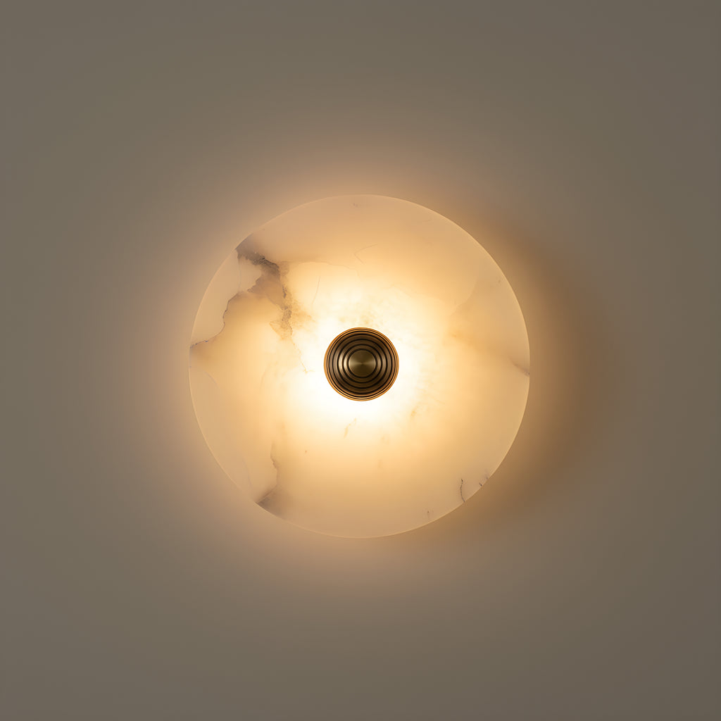 Clérachic alabaster disc wall light lit on a warm greige wall, its veining glowing through the stone around the ribbed brass boss at the centre