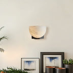 A Clérachic alabaster wall light on a cream wall above framed watercolour prints and small potted succulents on a shelf.