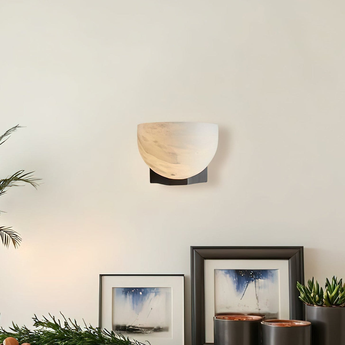 A Clérachic alabaster wall light on a cream wall above framed watercolour prints and small potted succulents on a shelf.