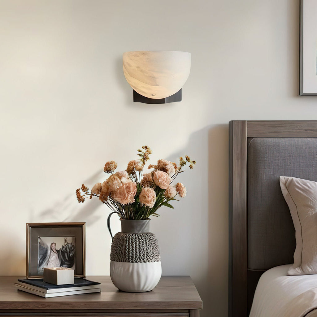 Above a bedside table with a patterned vase of peach flowers, this Clérachic alabaster wall light has a dark bracket against a cream wall.