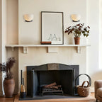Two Clérachic alabaster wall lights hang either side of a framed print above a fireplace mantel, with a dried-flower vase on the hearth.
