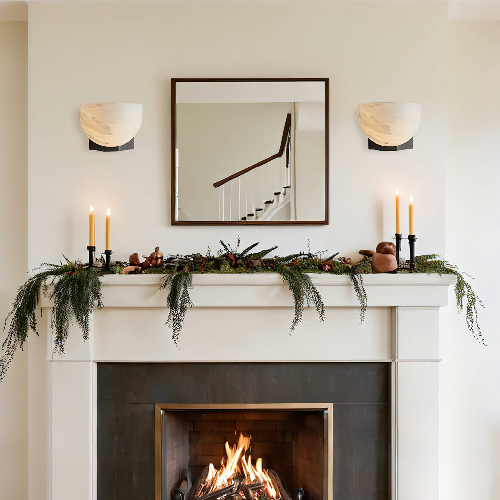 Either side of a square mirror, two Clérachic alabaster wall lights sit above a mantel dressed with greenery and tall candles.