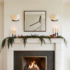 Either side of a square mirror, two Clérachic alabaster wall lights sit above a mantel dressed with greenery and tall candles.