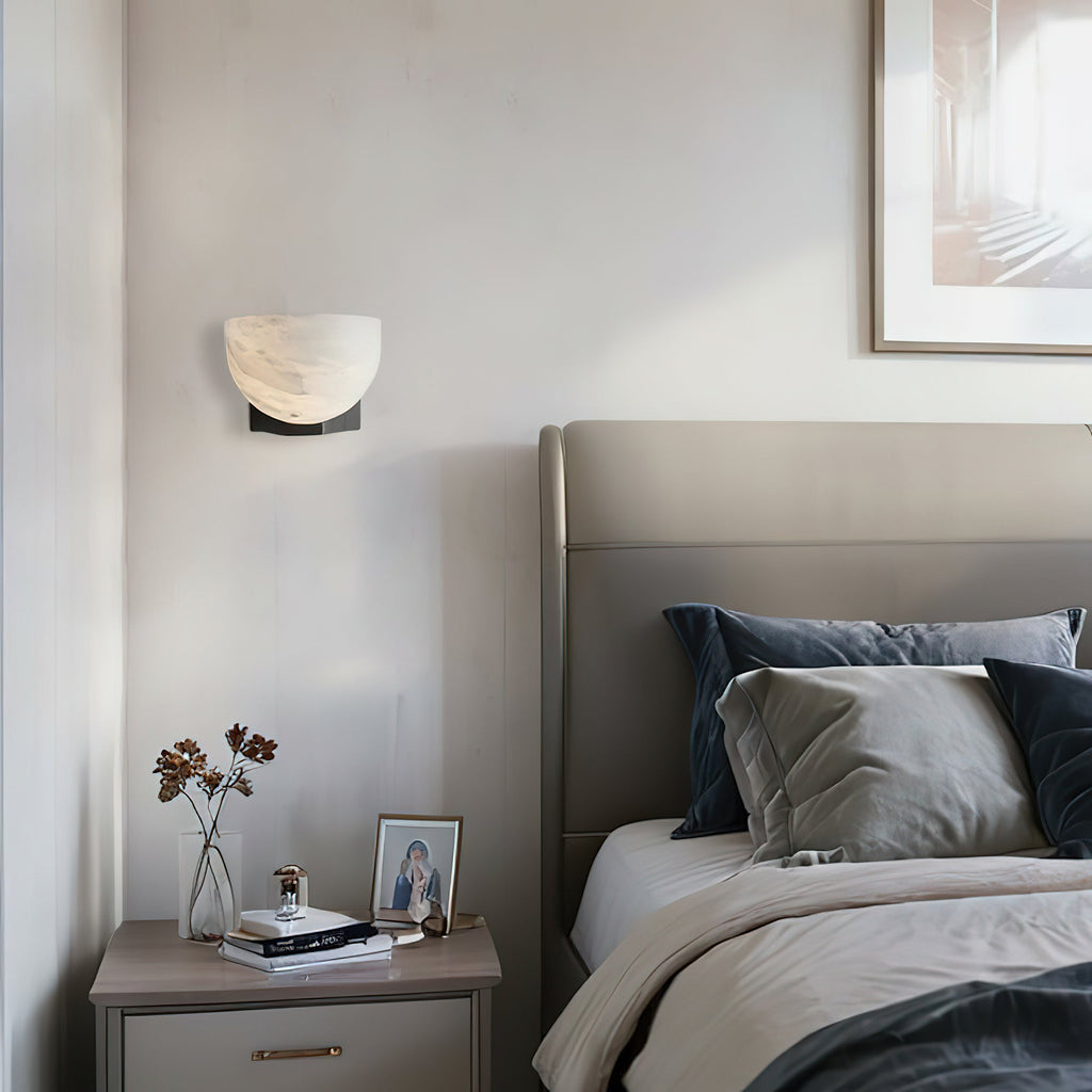 This Clérachic alabaster wall light with a dark bracket hangs above a bedside table and a grey headboard with navy cushions.
