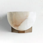 This Clérachic alabaster wall light unlit on a white wall, showing tan and grey veining in the stone above a brass bracket.