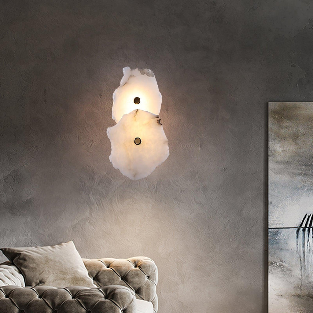 Clérachic's two-head alabaster wall lamp glowing softly on a textured grey wall beside a tufted sofa.