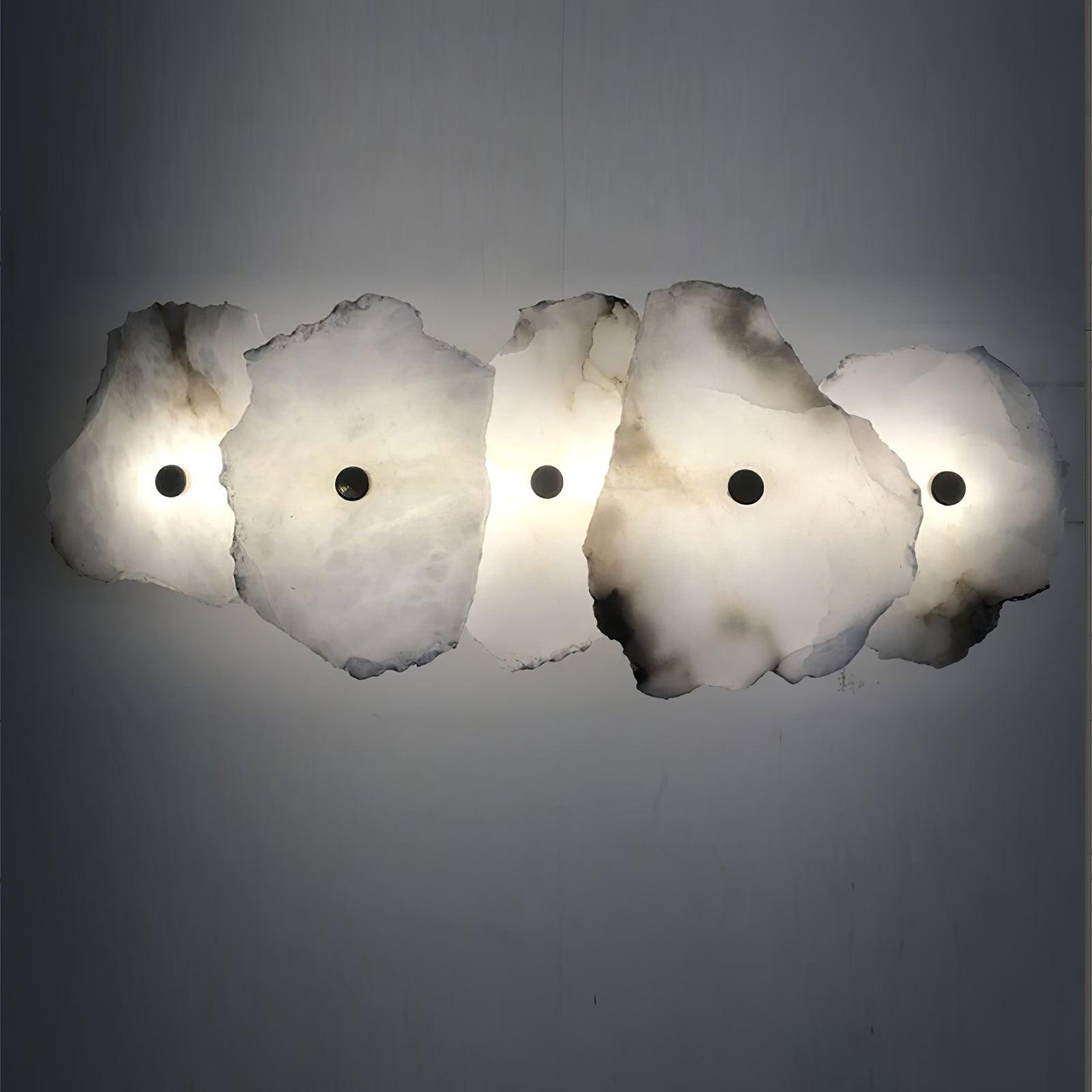 The Clérachic five-head alabaster sconce spans a grey wall, its stone shades glowing amber where light passes through.