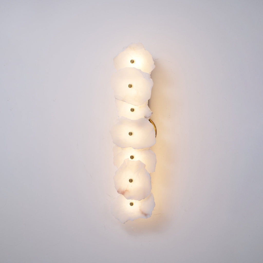 Clérachic's six-head alabaster lamp glowing softly on a plain wall, with the brass backplate visible at its edge.