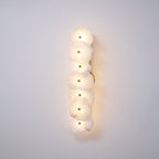 Clérachic's six-head alabaster lamp glowing softly on a plain wall, with the brass backplate visible at its edge.