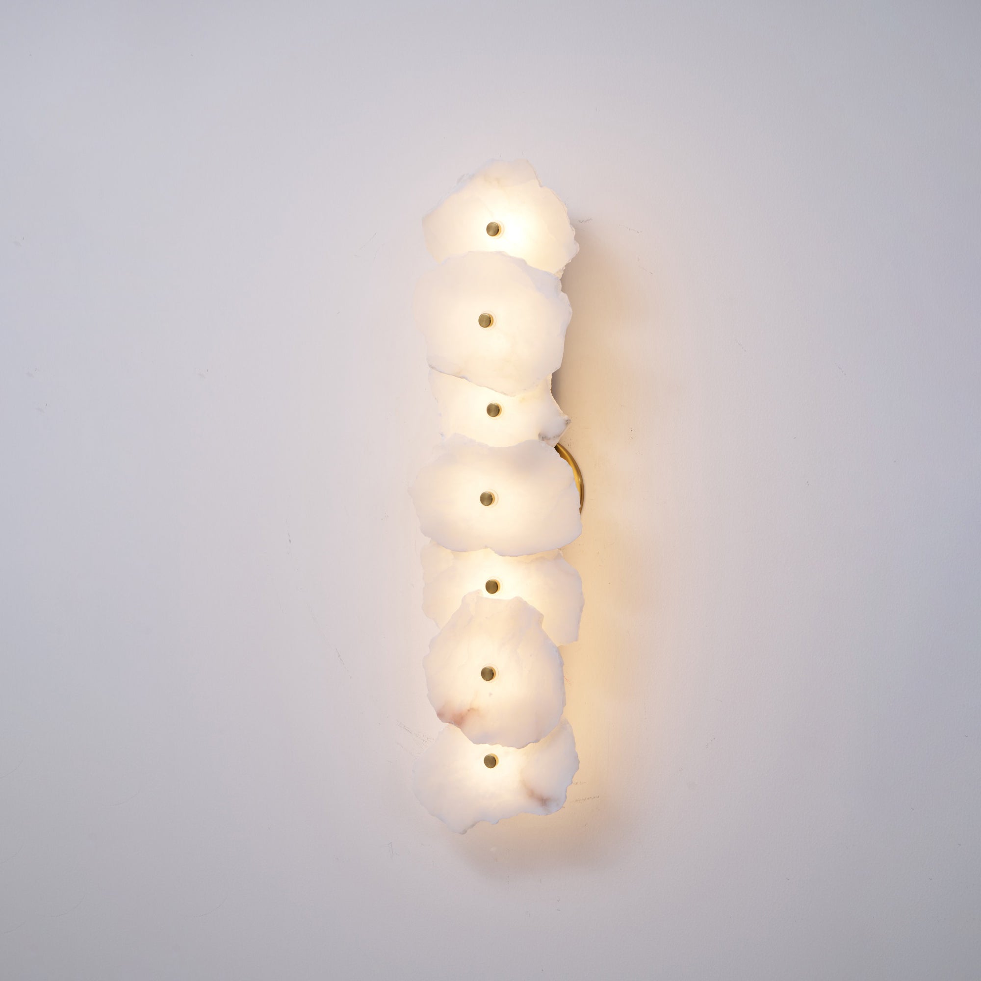 Clérachic's six-head alabaster lamp glowing softly on a plain wall, with the brass backplate visible at its edge.