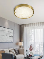Round Clérachic alabaster flush ceiling light glowing above a living room sofa, artwork and a sheer-curtained window.
