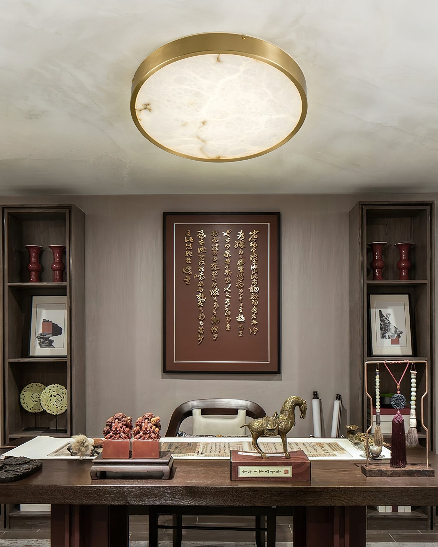 Round Clérachic alabaster flush ceiling light over a study desk, its stone face lit above framed calligraphy and a bronze horse.