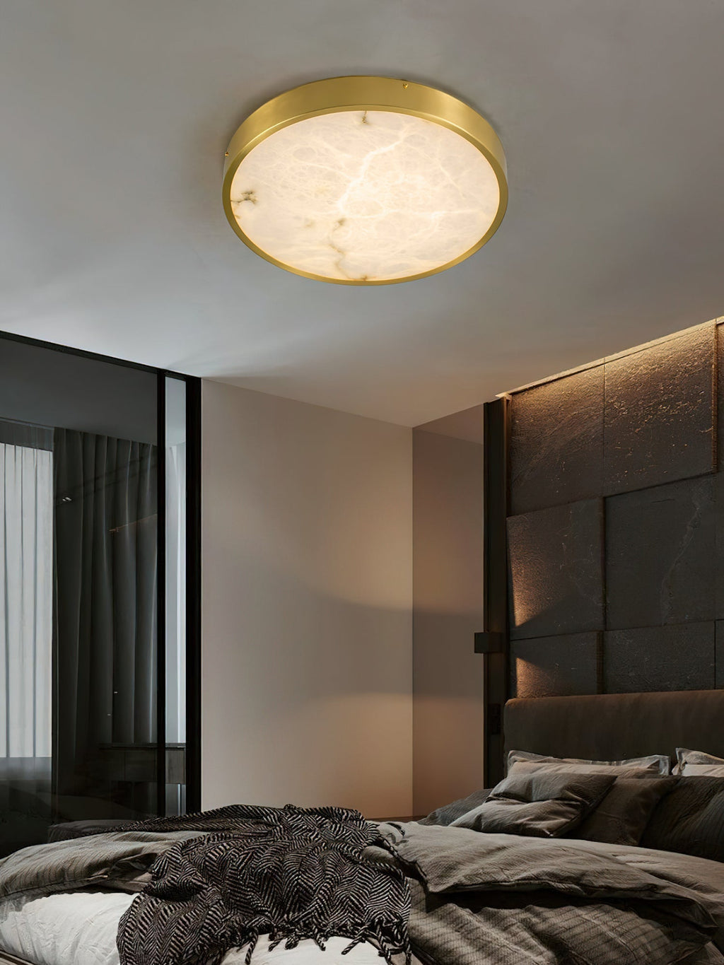 Clérachic alabaster flush ceiling light lighting a dark bedroom, the round stone face soft against a stone headboard wall.