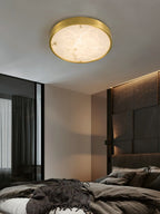 Clérachic alabaster flush ceiling light lighting a dark bedroom, the round stone face soft against a stone headboard wall.