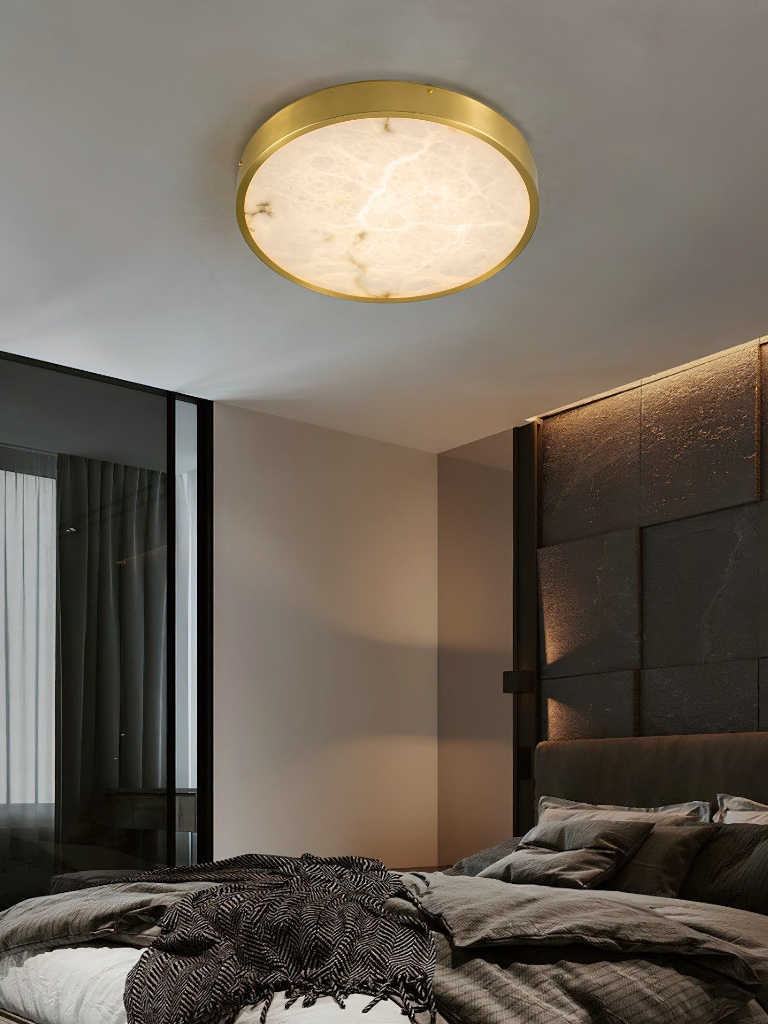 Clérachic alabaster flush ceiling light lighting a dark bedroom, the round stone face soft against a stone headboard wall.