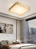 Square Clérachic alabaster flush ceiling light lit in a bedroom, between two abstract prints and a city window.